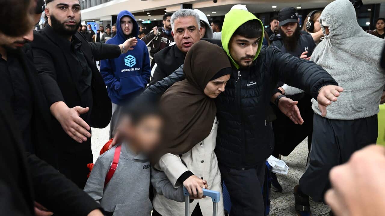 A woman and child are guided through a very crowded airport by a hooded man.