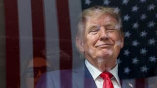 US President Donald Trump in a dark suit with a bright red tie standing in front of an American flag. The image is photographed through reflective glass, creating a layered effect with flag stripes and stars overlaid across the person's face and body. A faint reflection of another person wearing dark glasses appears in the lower left of the frame.