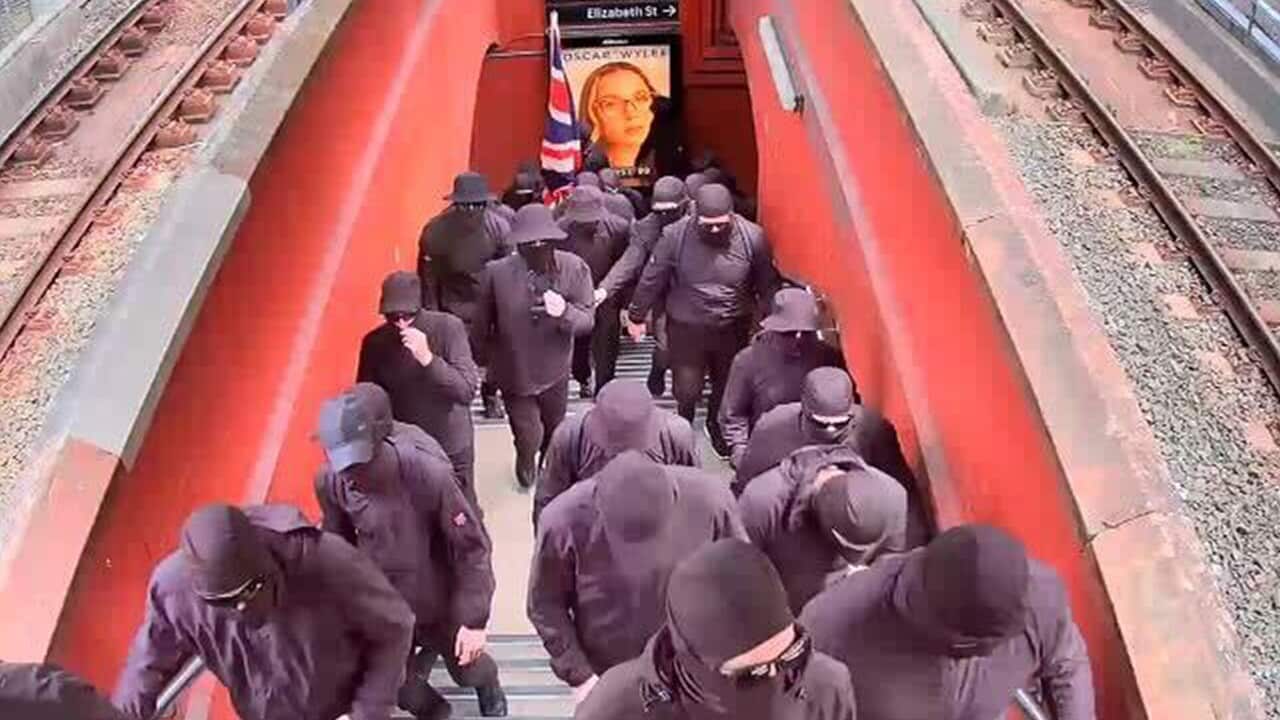 A large group of people wearing black clothes and face coverings walk up a set of stairs at a train station.