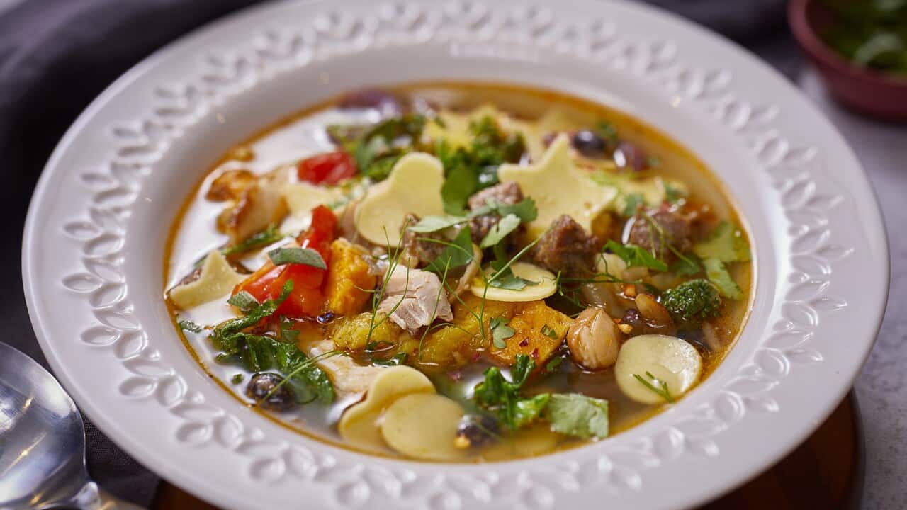 A wide white soup bowl with a textured floral pattern on the rim holds a birght soup, full of vegetables, meat and herbs.