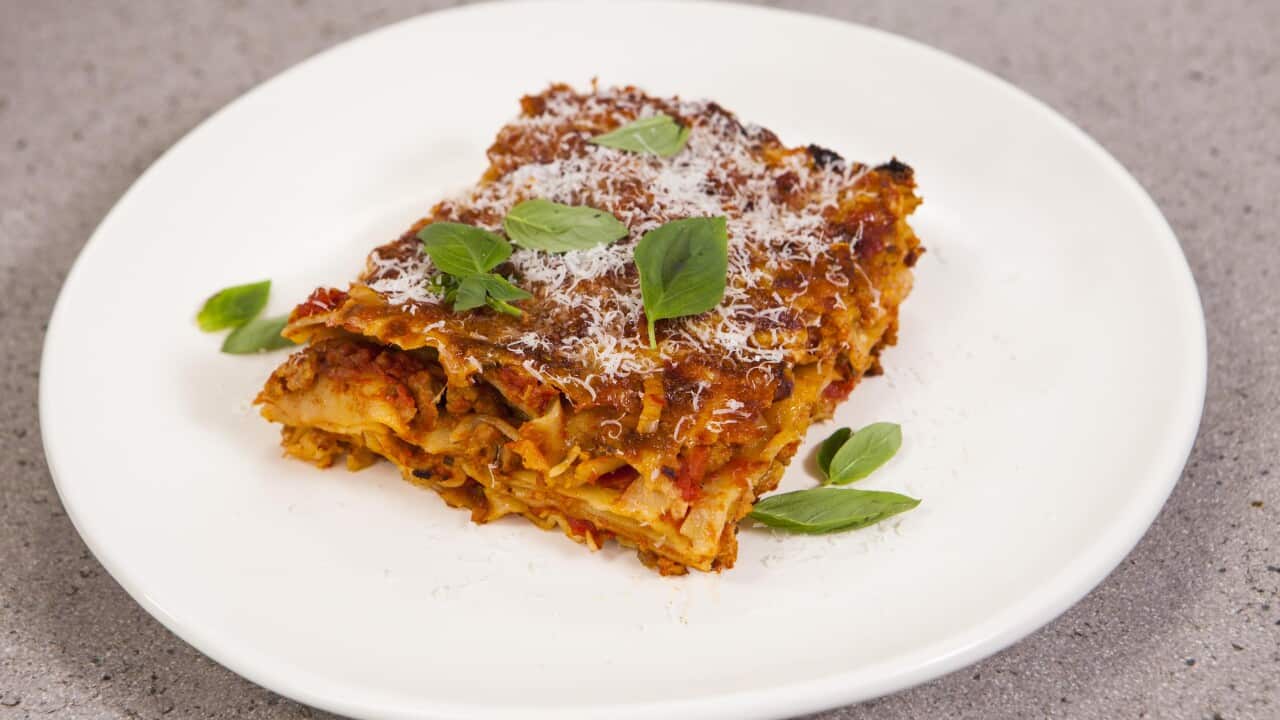 A rectangular piece of lasagne sits on a round white plate, scattered with a few basil leaves.
