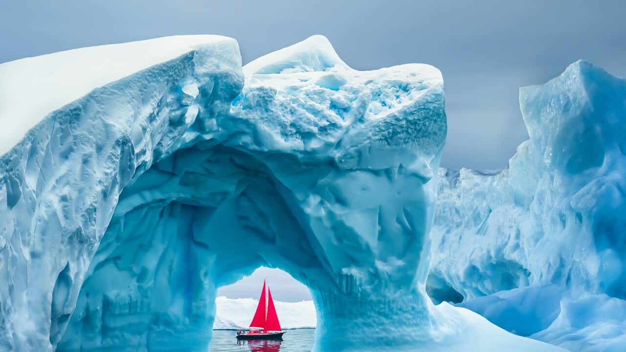 Red sailboat sailing under a majestic iceberg arch on sunny blue Artic Ocean in Greenland, Ilulissat Icefjord, Ilulissat, Disko Bay, Unesco World Heritage Site