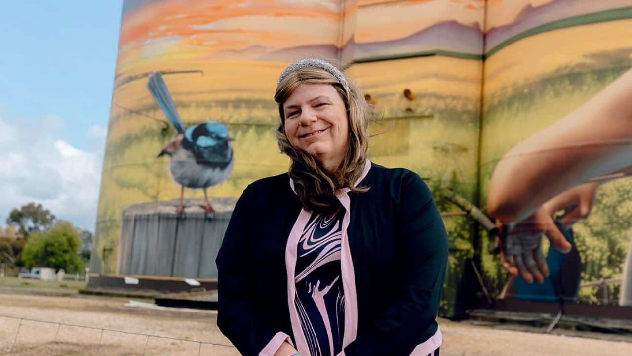 A woman standing in front of a rural backdrop with a mural of a blue bird, a child and a country landscape painted onto two water towers behind her.