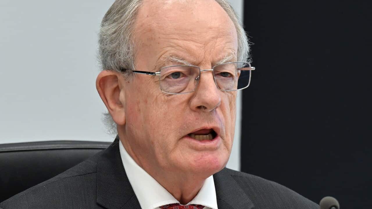A man wearing a suit, tie, and glasses is seated and speaking in front of a microphone.