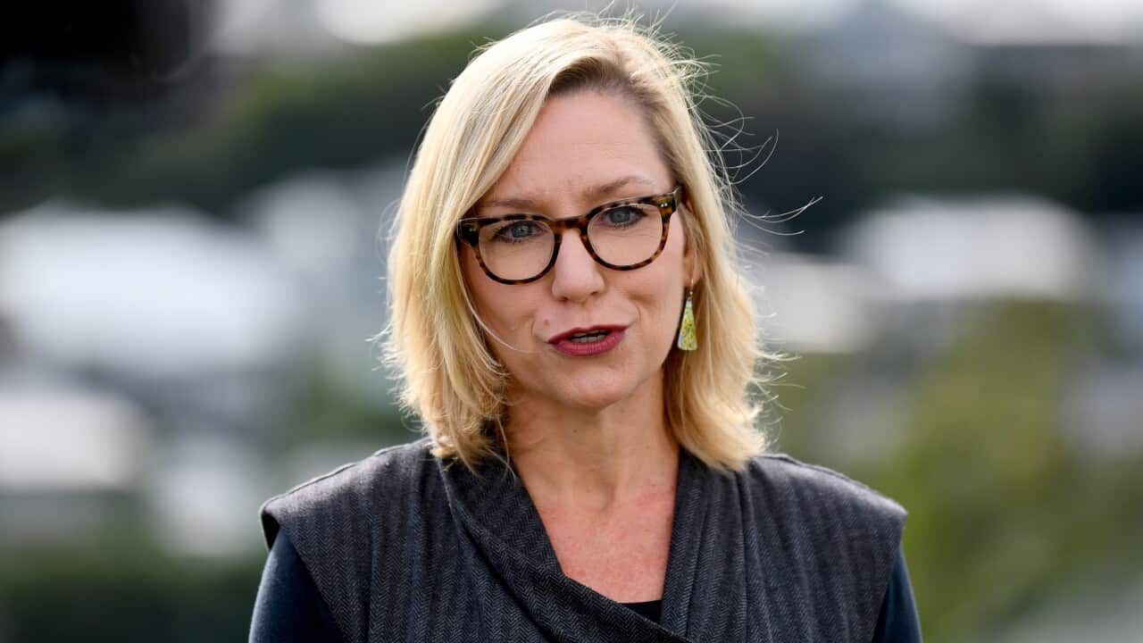 A blonde woman with black glasses and red lipstick speaking to a camera