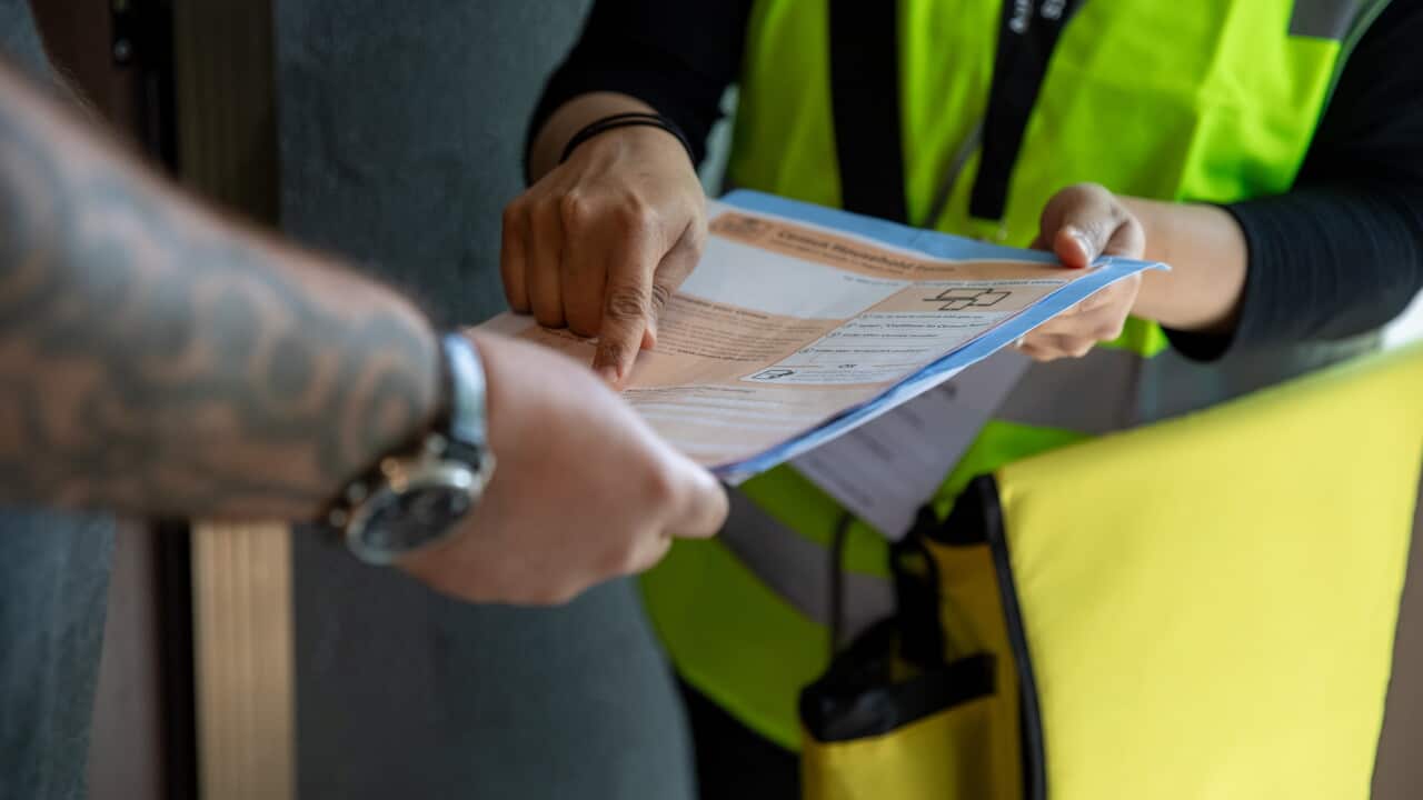 A woman wearing a fluorescent yellow vest hands a man an orange Census form. He wears a watch and has tattoos on his forearm.