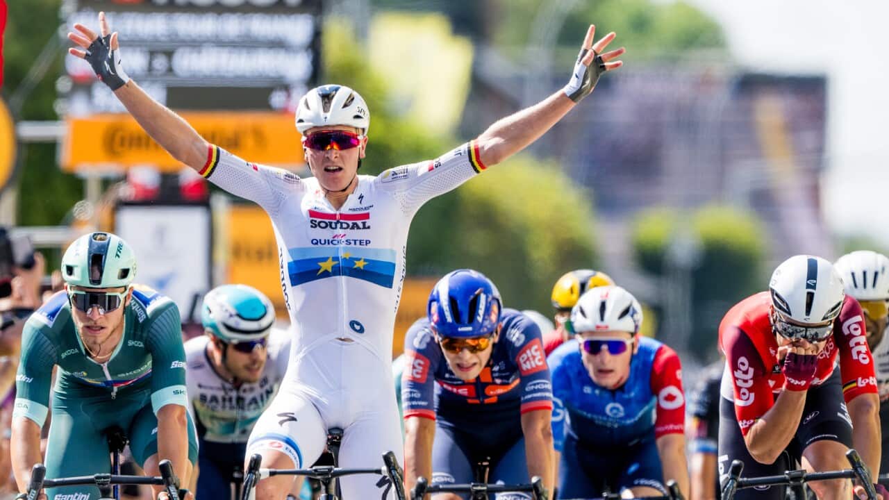 Group of cyclists at the finish line of the race. The central rider, wearing a white jersey, has both arms raised in victory.