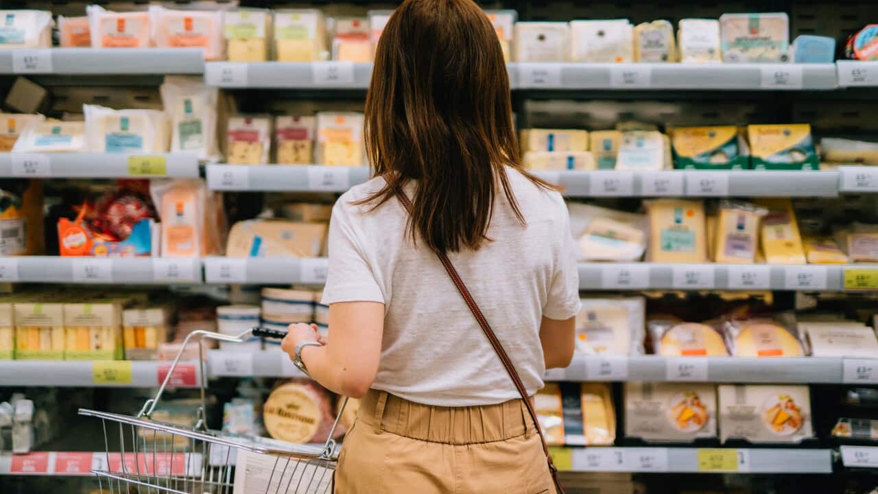 Rear view of young woman carrying shopping basket, choosing cheese, standing in front of produce aisle in supermarket