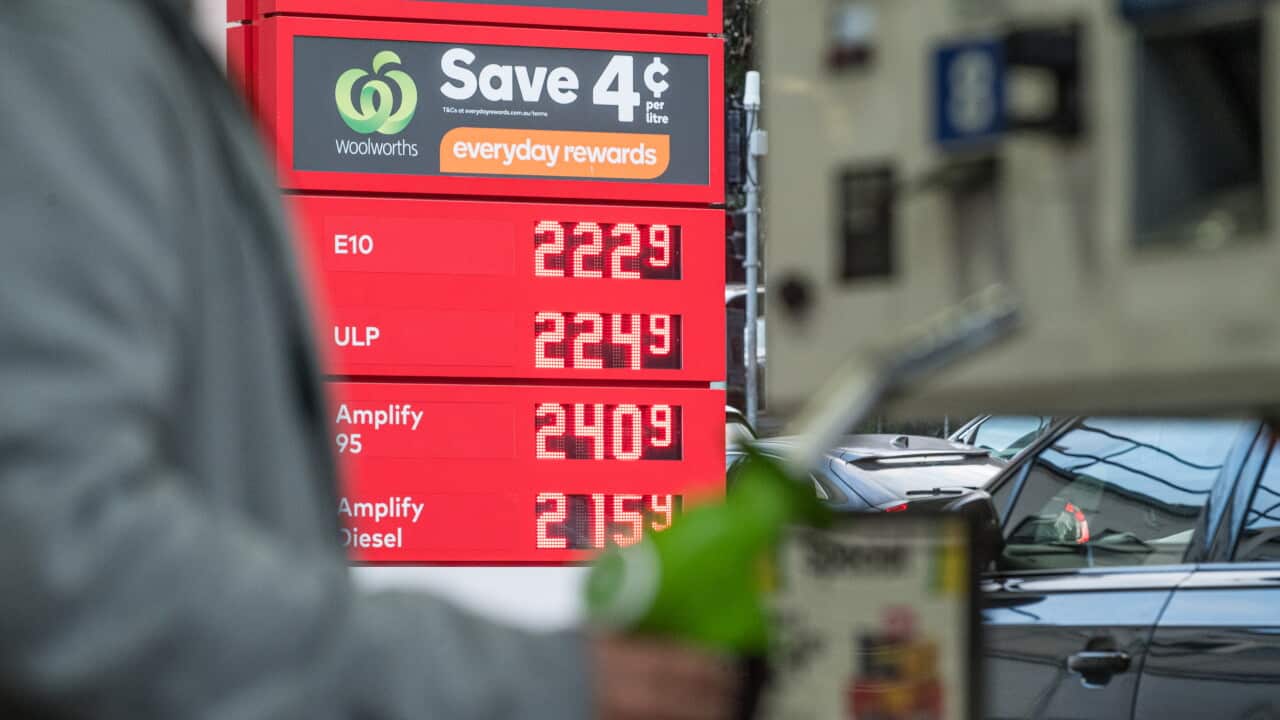 A red digital price sign at a petrol station, showing fuel prices in cents per litre. A blurry figure holds a green fuel pump nozzle in the foreground.