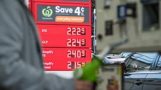 A red digital price sign at a petrol station, showing fuel prices in cents per litre. A blurry figure holds a green fuel pump nozzle in the foreground.