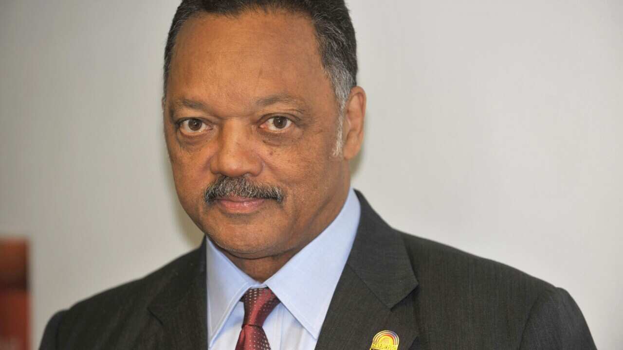 Jesse Jackson, wearing a grey suit, light blue shirt and red tie, smiling at the camera.