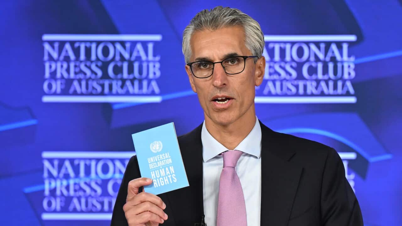 An image of Australian Human Rights Commission president Hugh de Kretser. He is an older man with greying hair and glasses. He wears a black blazer, blue shirt, and mauve tie, and holds a pamphlet in his hand.