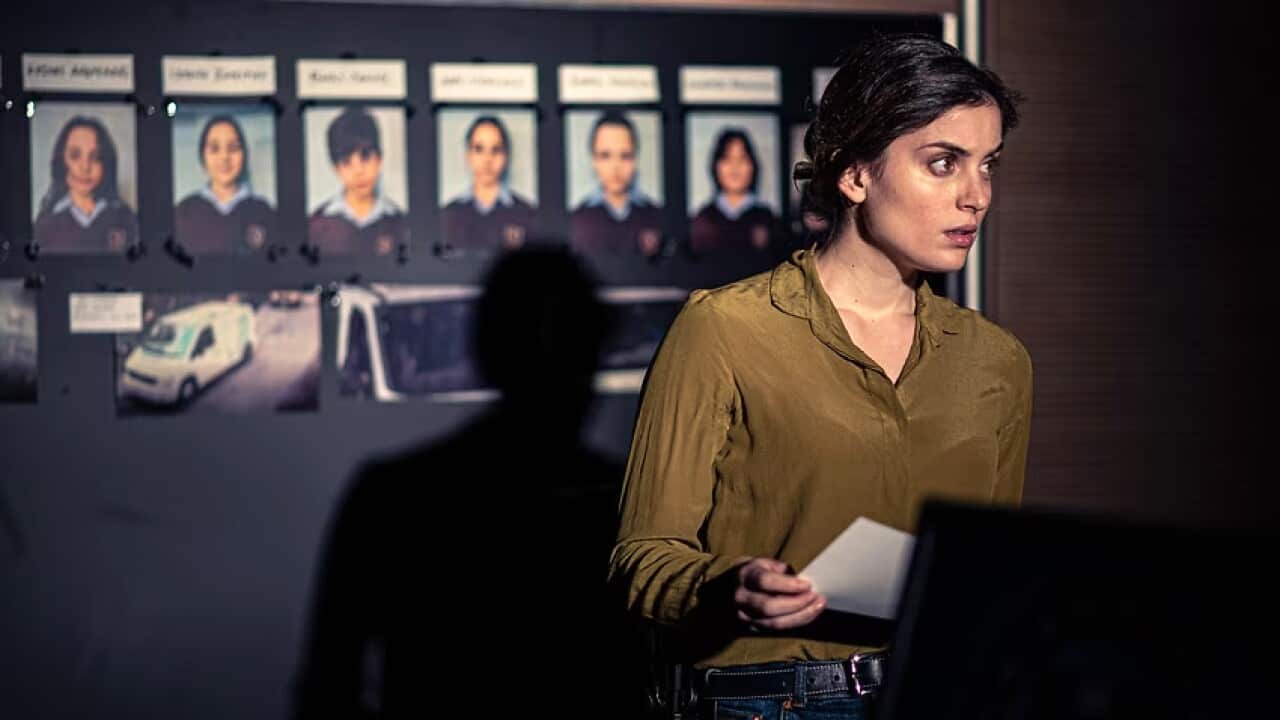 A woman holds a piece of paper, standing in front of a board with pictures of a crime investigation.