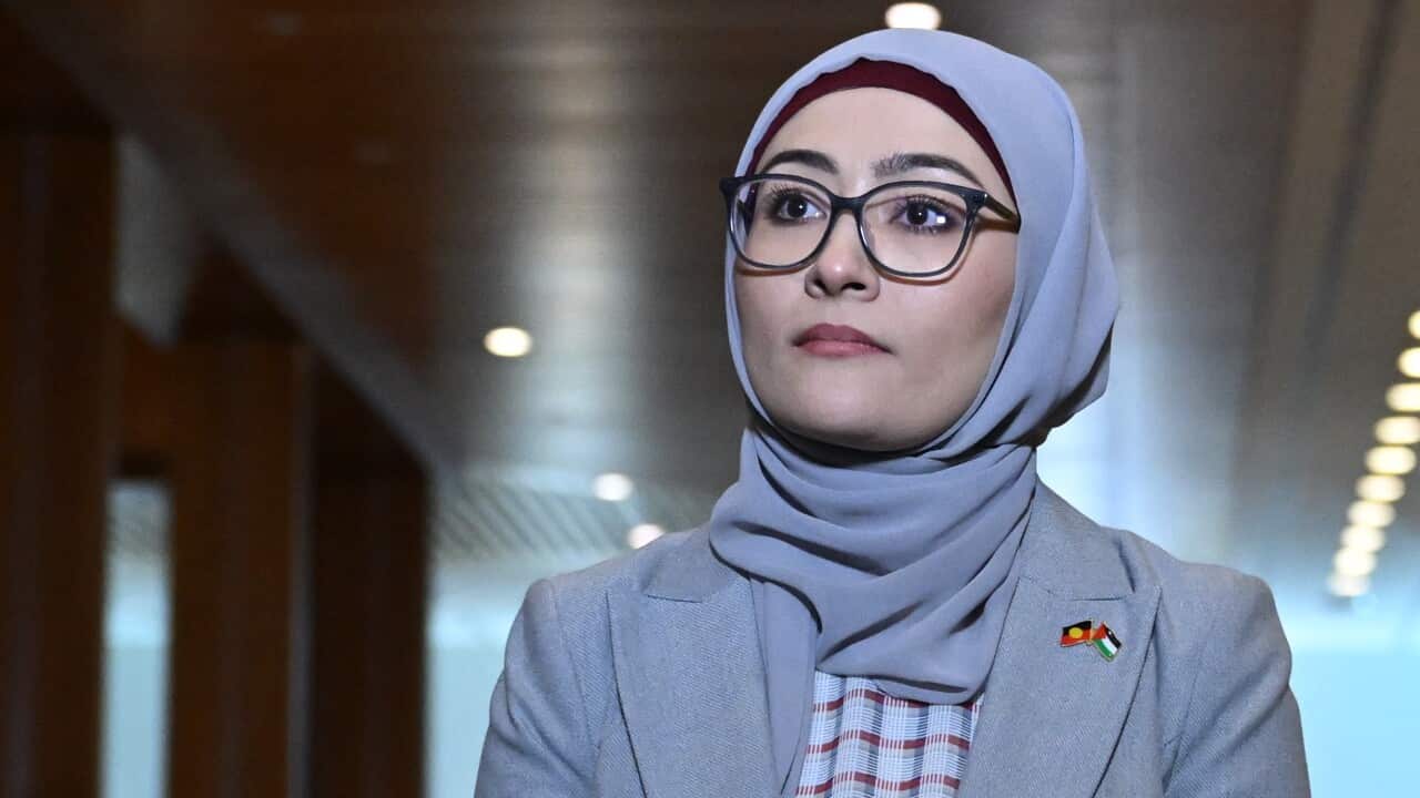 A woman standing in a corridor. She is wearing pins that show overlapping Aboriginal and Palestinian flags.