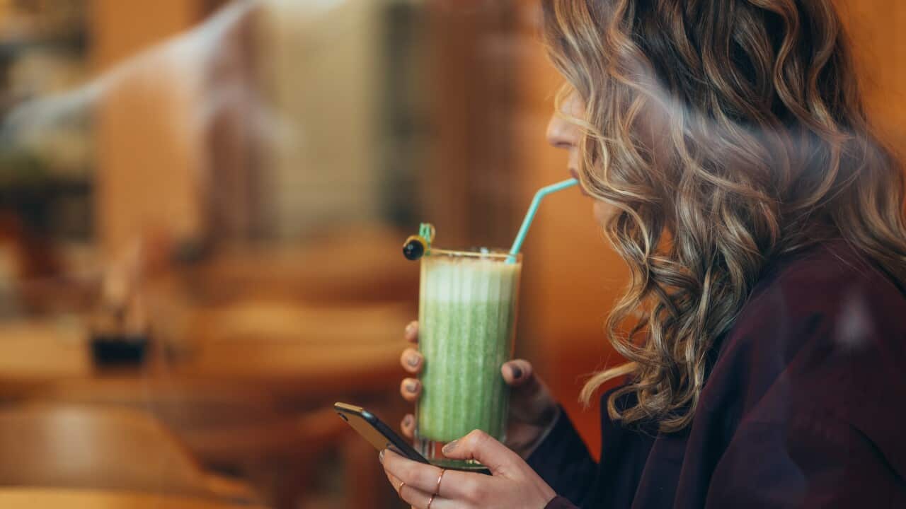A woman drinking a glass of matcha, holding it in one hand and her phone in the other.
