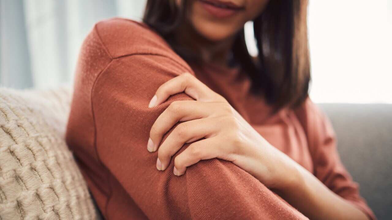 Close up of a young woman holding her arm and suffering with pain while sitting in the living room at home. Mixed race woman holding her arm in pain due to injury or arthritis
