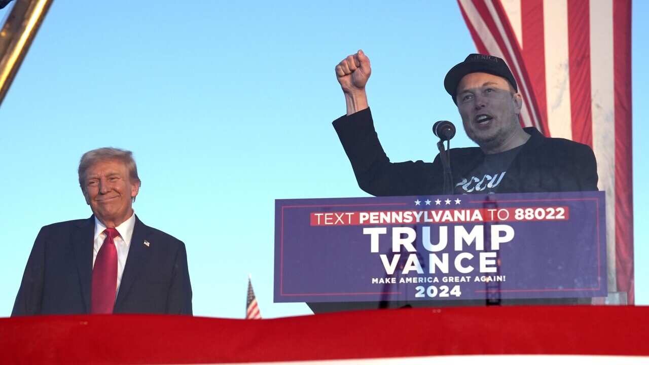 A woman with short grey hair, wearing a black jacket and glasses, looks on.