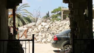 View through a shattered building opening onto a large pile of rubble and twisted rebar, with a damaged car, palm tree and damaged structures visible beyond.
