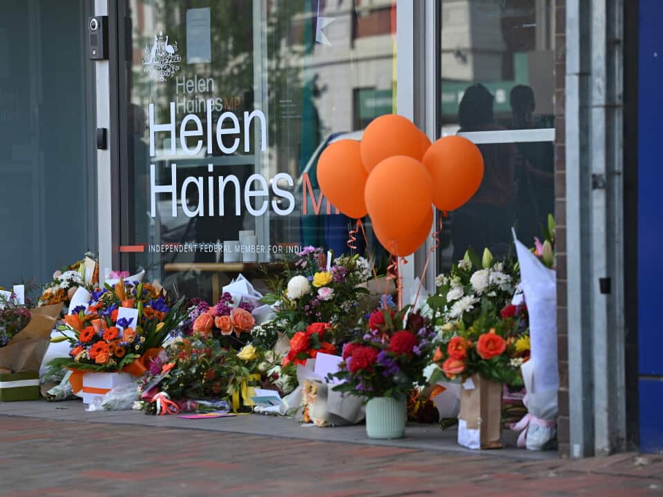 A large pile of flowers with a bunch of orange balloons sits against a window with a sticker reading "Helen Haines MP".