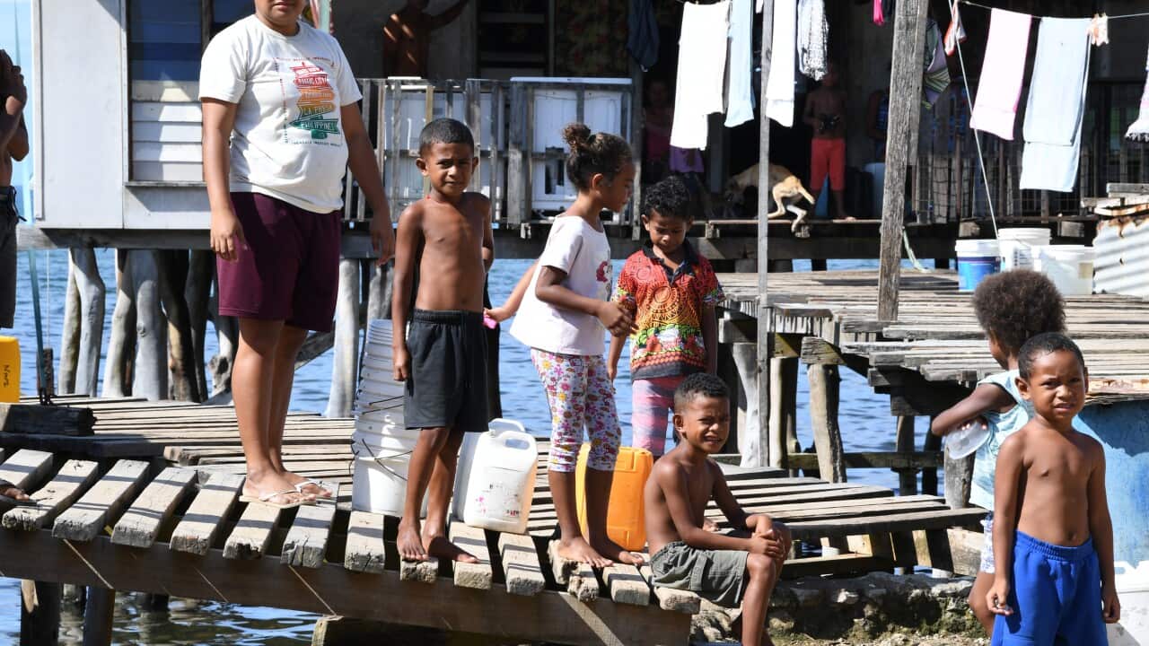 Children standing on a pier, with a house behind them on stilts.
