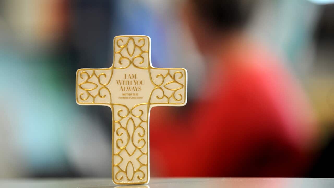 A small cross with gold filagree. A bible verse is inscribed on it. A person in a red shirt sits in the background, blurred out.