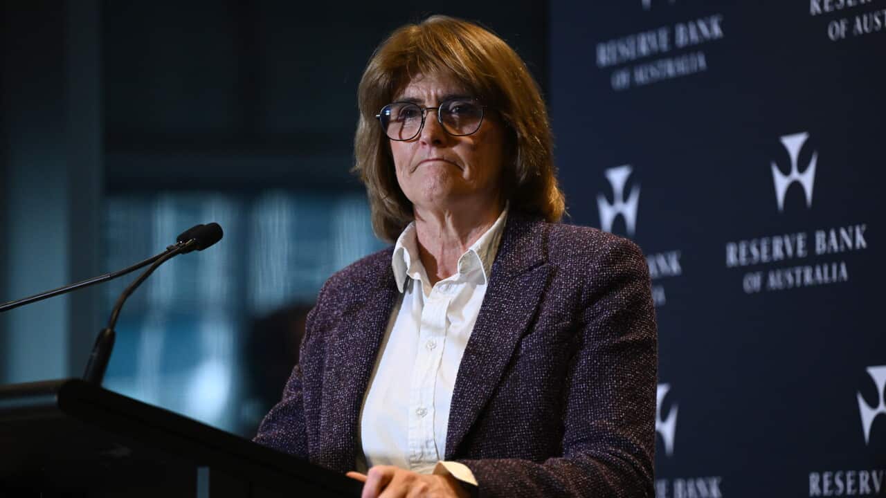 Woman wearing glasses and a purple blazer stands at a lectern before a Reserve Bank of Australia backdrop, speaking into a microphone during a public address.