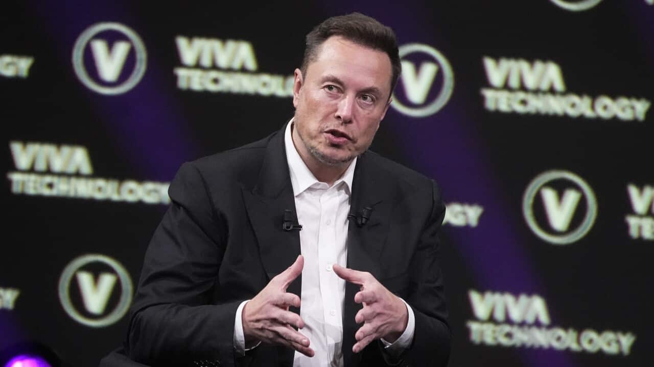 Elon Musk in a black suit and white shirt sitting down speaking and gesticulating with his hands.