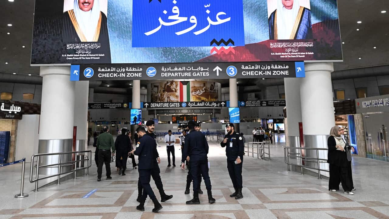 Security officials stand beneath a screen displaying photos of two Emirs at an airport terminal.