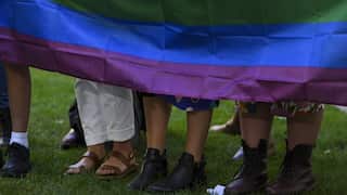 A collection of people's feet in shoes, standing on grass. A rainbow pride flag drapes to their knees.