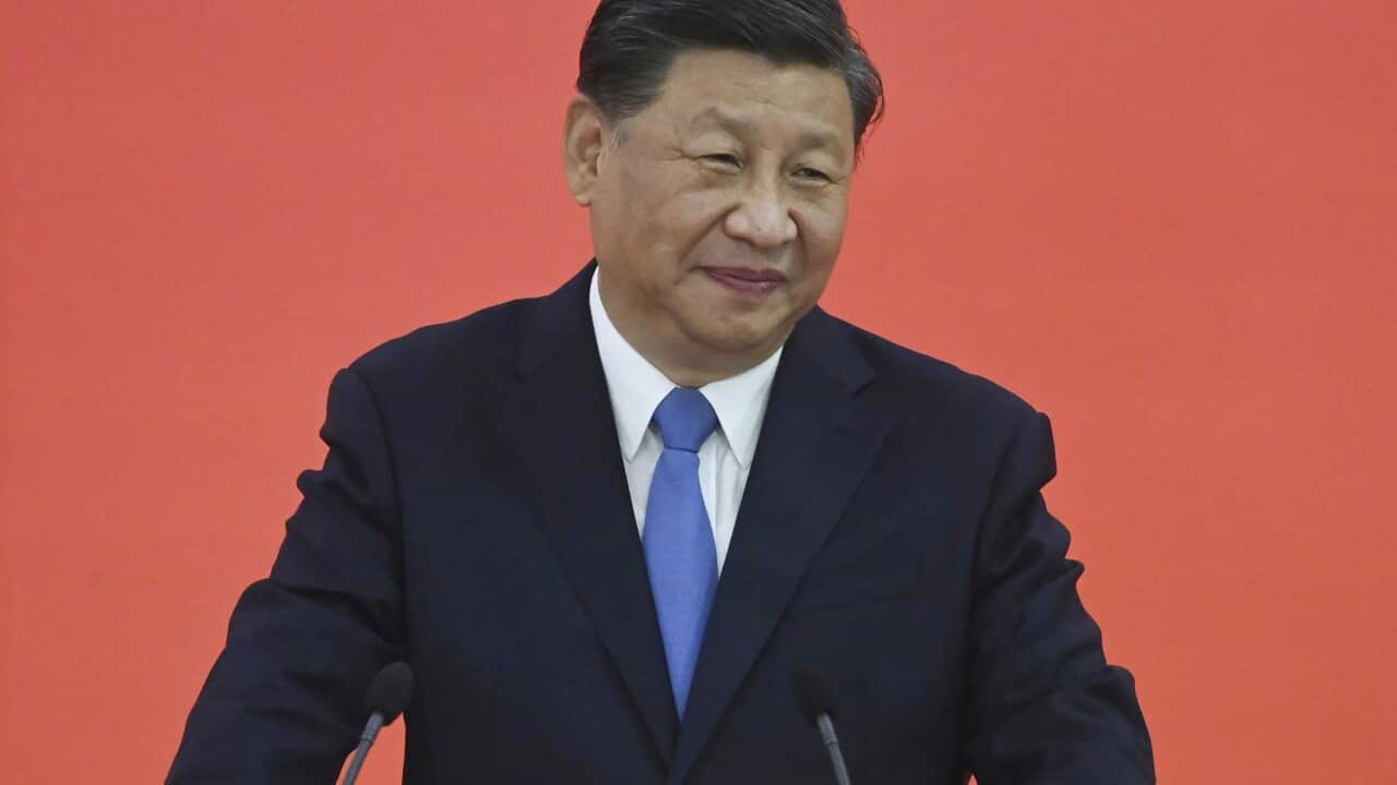 Xi Jinping stands at a podium in a suit and blue tie, with a small smile.