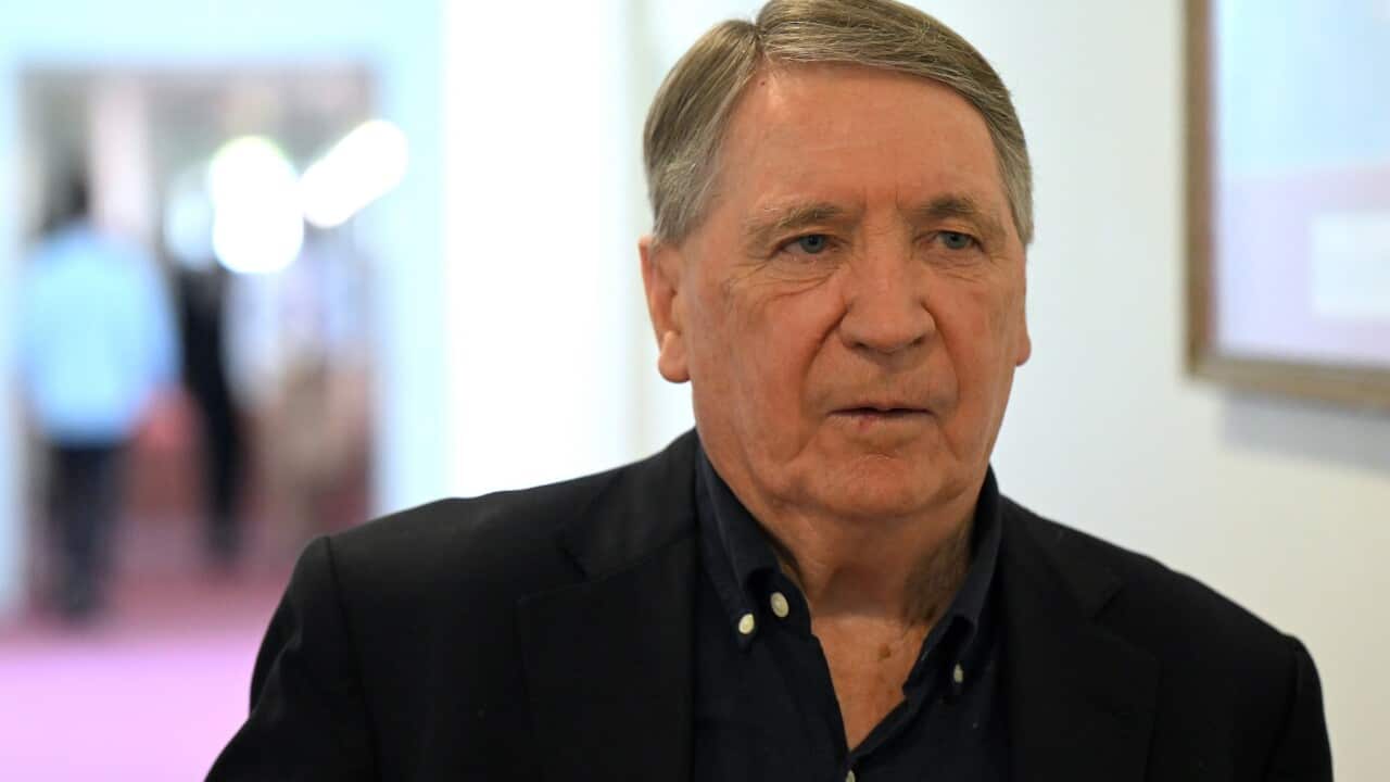 A man in a dark blue shirt and black blazer, standing in a hallway.
