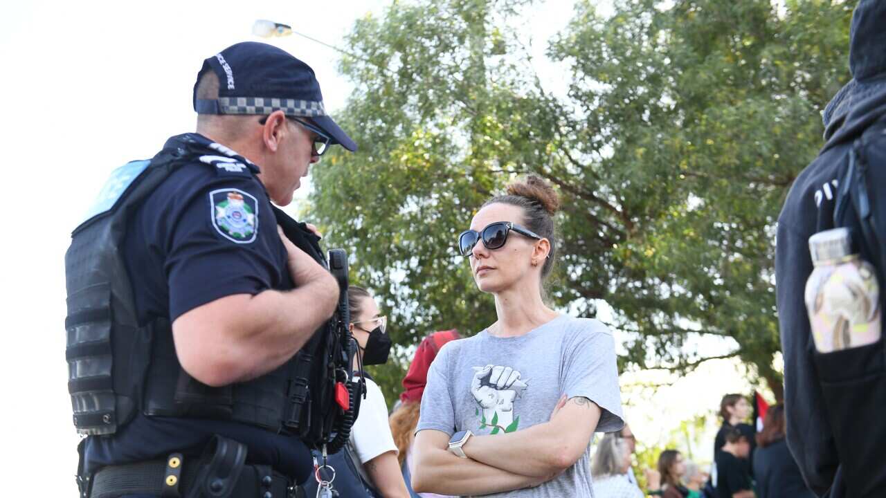A woman wearing a grey T-shirt and sunglasses stands with her arms folded in front of a male police officer.