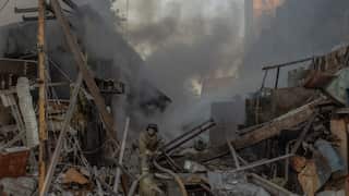 A firefighter, covered in dirt, is carrying a large hose into the rubble of a bombed residential building.