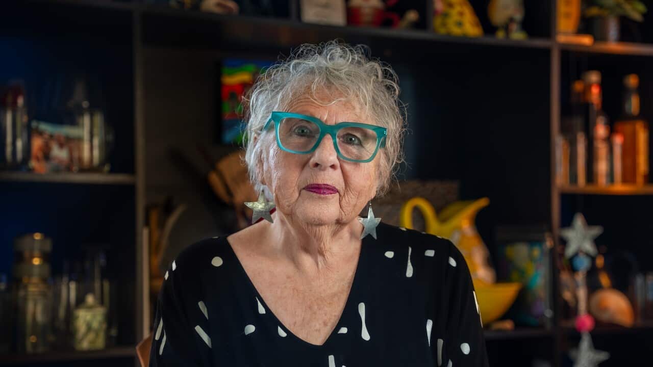 A woman in green glasses sits in front of a bookshelf.
