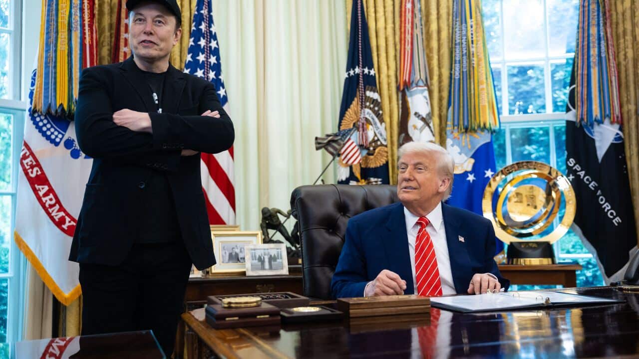 Elon Musk, wearing all black and a black cap and standing with his arms crossed, stands next to Donald Trump, who is wearing a dark suit and red tie and sitting at a desk in the Oval Office.