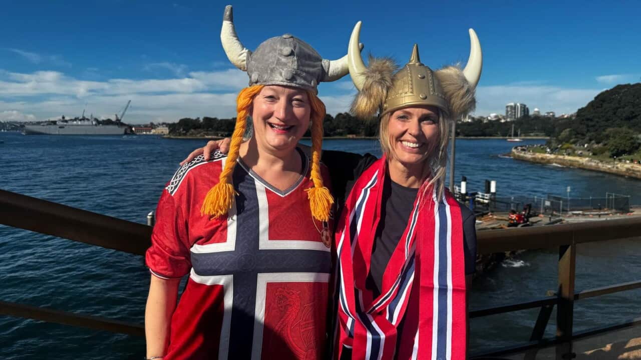 Two white women stand draped in Norwegian flags and Viking helmets in front of Sydney Harbour.
