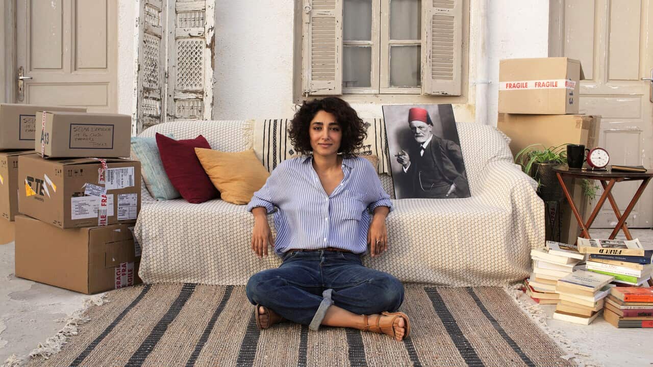 A woman in blue jeans and a blue shirt sits on a rug on the floor, in front of a couch. Packing boxes are stacked to either side of the couch. Several piles of books sit on one side.