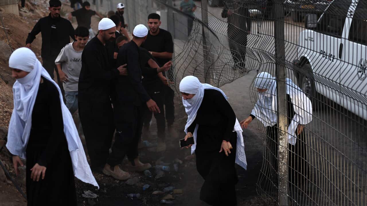 People, including women in traditional dress, pass through a bent fence while others gather nearby in a dusty area next to a police vehicle.