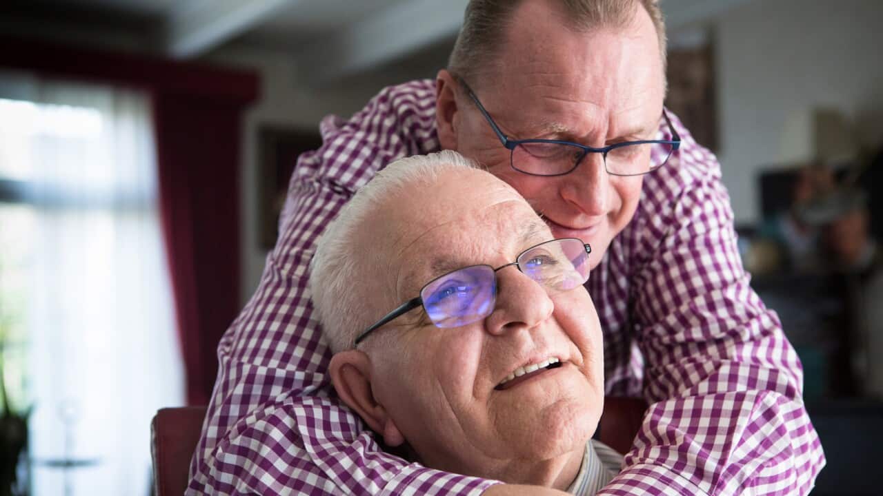 One man is sitting in a comfortable chair, the other man is standing behind him. He has is arms around the sitting man.a homosexual couple, almost 25 years together in and around their house.twenty years ago they were the first homosexual pair that got ma