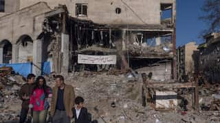 A family of four walks past the massive pile of rubble and exposed structural beams of a heavily destroyed building featuring a banner with Arabic script.