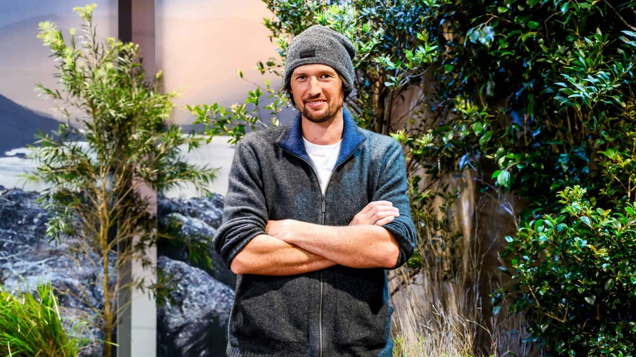 A man wearing a jumper and beanie smiling at a camera with plants in the background