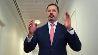 Ed Husic, a middle aged man with brown hair and a greying beard, dressed in a blue suit with red tie, stands in a white hallway.