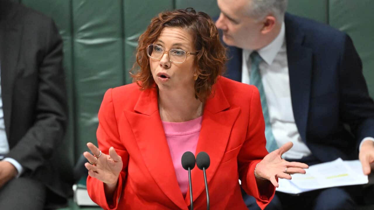 A woman in an orange blazer speaks into a microphone with her hands raised.