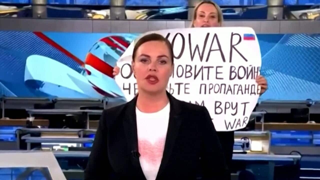 A woman is seen standing behind a news anchor holding a sign that says "no war".