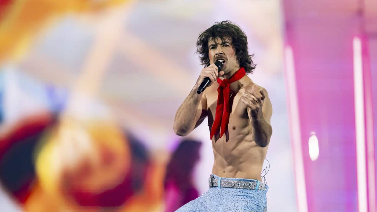 A man wearing light blue pants and a red scarf singing with a microphone in hand.