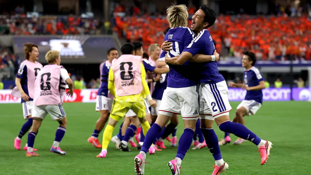Two players wearing blue football shirts and white shorts hug as they jump together on a football field. Other players celebrate in the background
