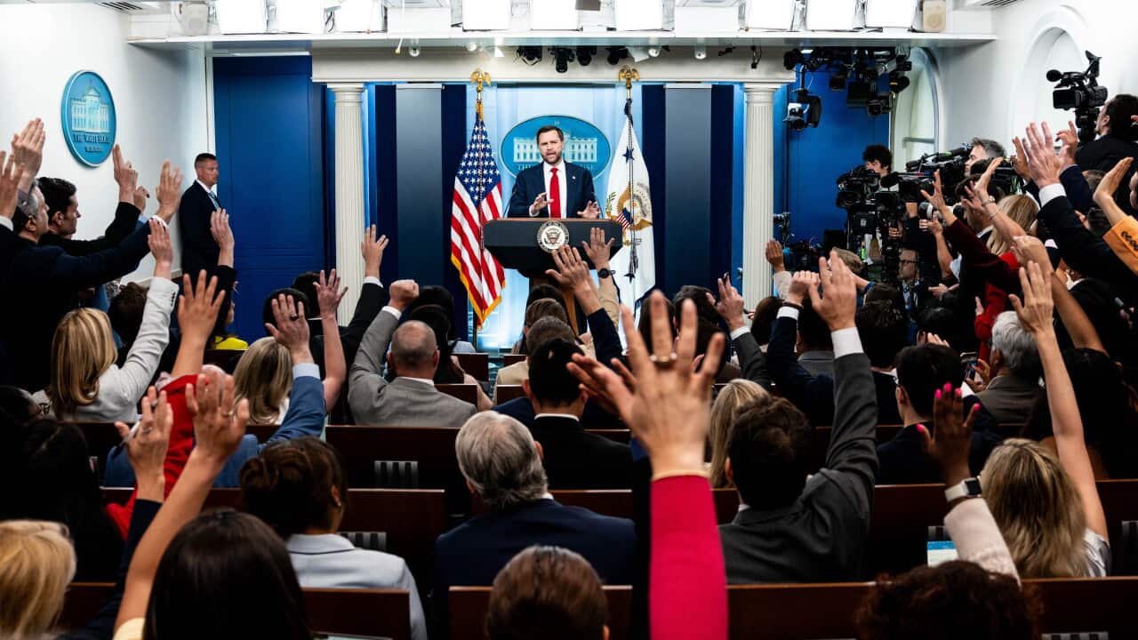 A man stands at a podium delivering a speech before a room of reporters with their hands raised.