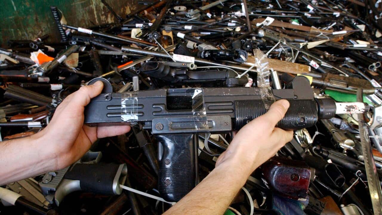 Man holding a gun above a stockpile of many more guns of different shapes and sizes.