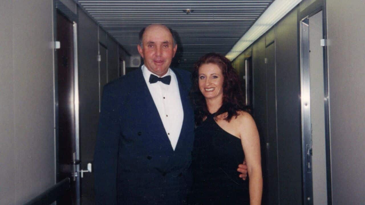 a film photo of a couple in formal wear standing in a dark hallway. The middle-aged man has his arm around the younger woman's waist