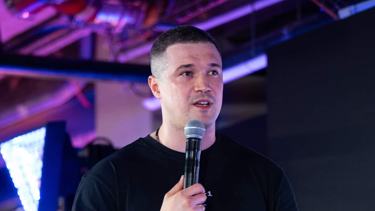 A man on a stage in a black T-shirt speaking into a microphone.